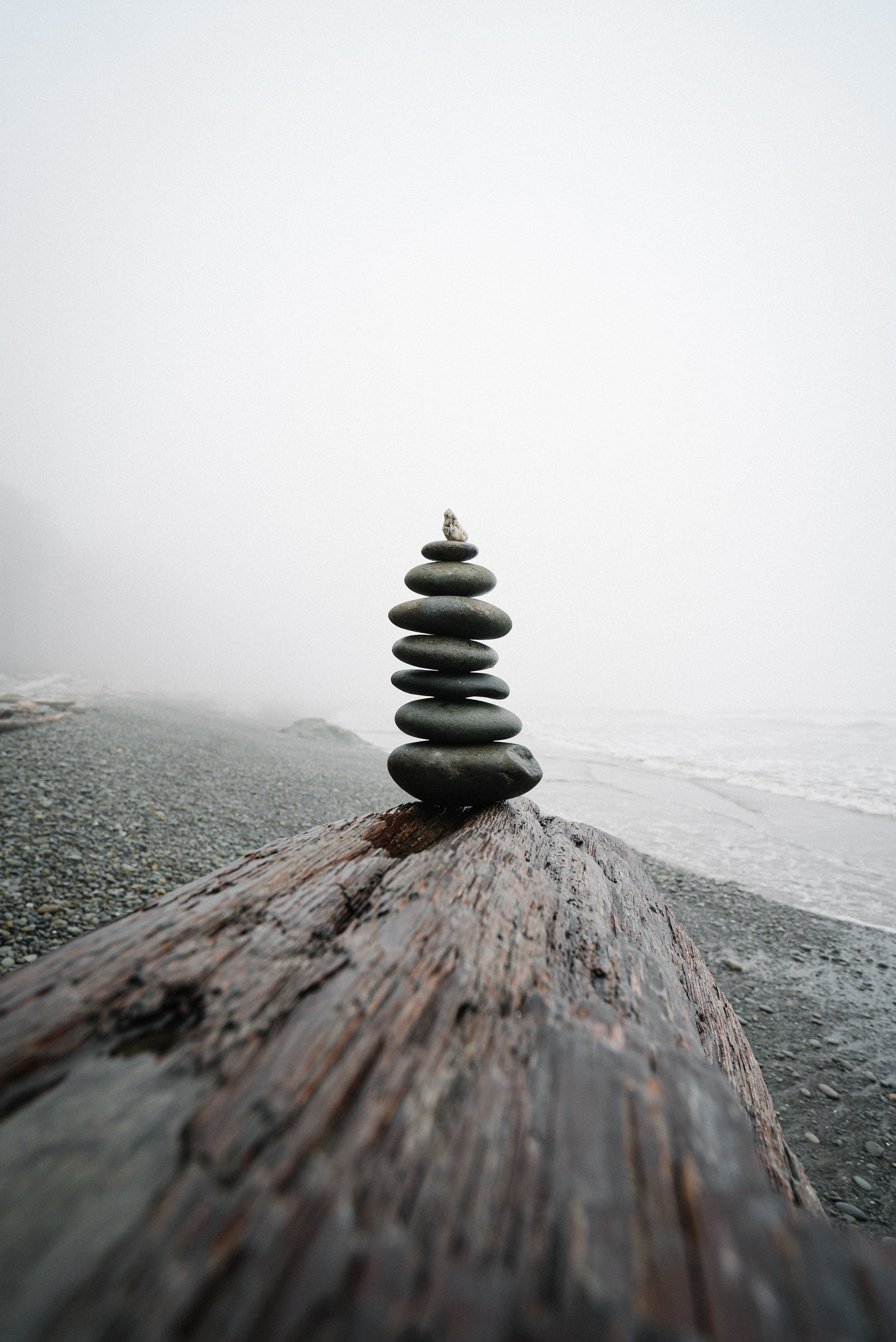 7 flache, dunkle Steine stehen aufeinander gestapelt 
                                                                                                                           auf einem Holzbrett am Strand vor dem Meer.