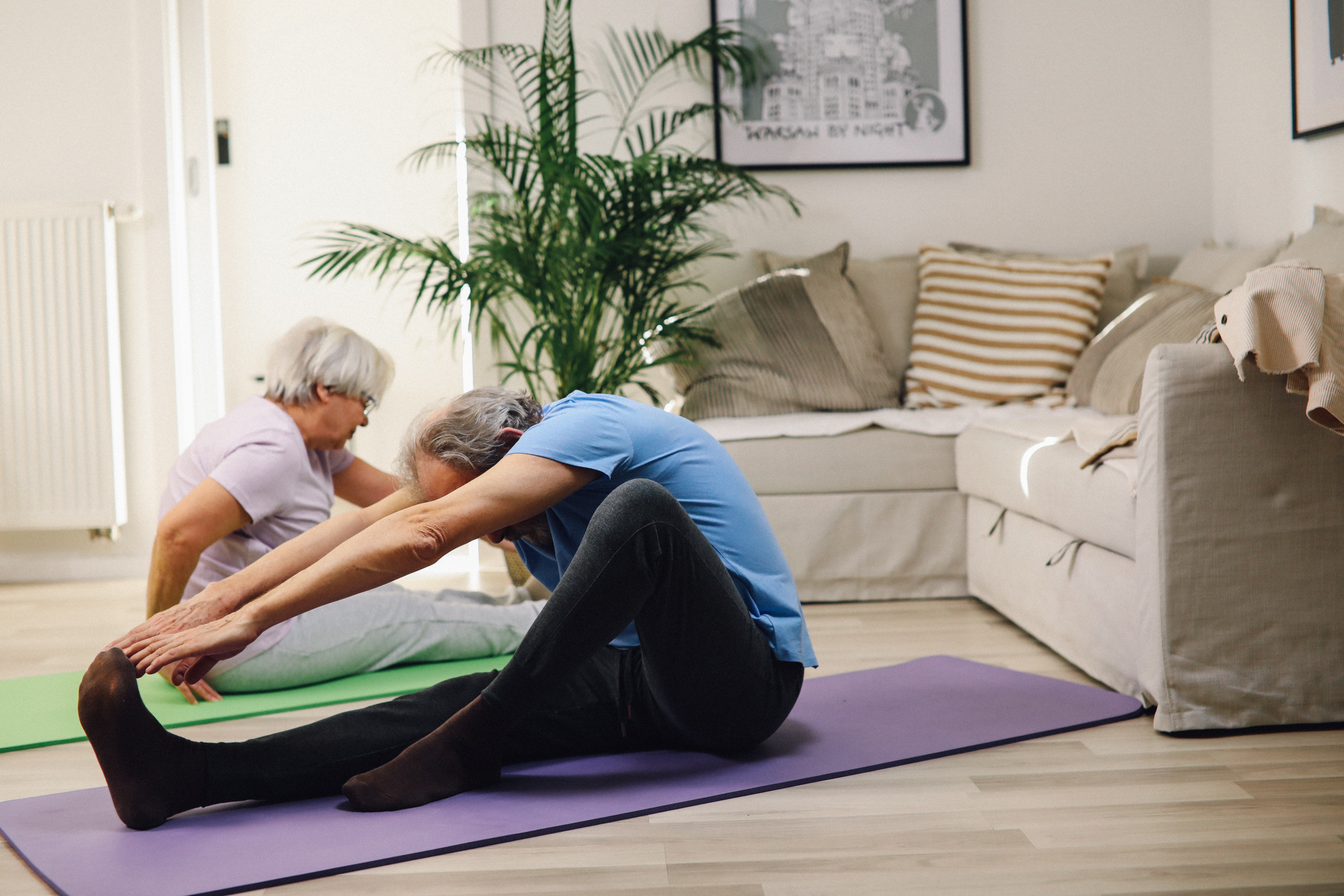 Ein Ehepaar sitzt gemiensam auf Yoga Matten im Wohnzimmer am Boden
                                                                                                                                    und dehnt sich. Sie bereiten sich auf ein Training vor.